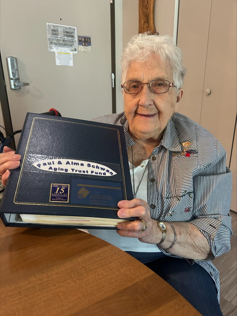 Elderly woman with white hair and glasses smiles while holding a large binder labeled Paul & Alma Schwarz Aging Trust Fund at a table, wearing a striped shirt and sitting in a cozy room.