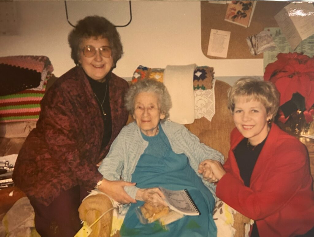 Three women, two standing and one seated in the middle, smile at the camera in a cozy living room. The seated woman holds hands with the others; colorful blankets and papers are visible in the background.