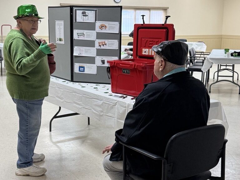 Two older adults are chatting near a display board and red coolers in a community room. One wears a green hat and sweater, the other a black cap and jacket. Tables and chairs are arranged around the room.