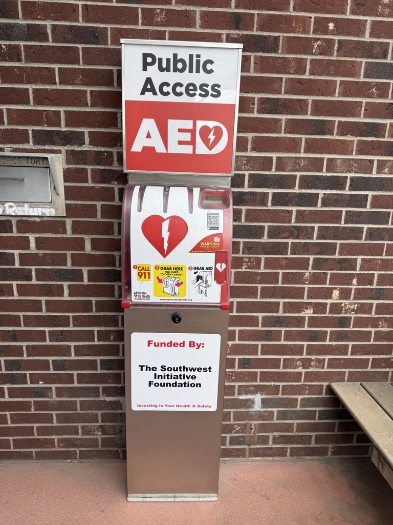 A public access AED station mounted on a brick wall, with clear signage and instructions, funded by The Southwest Initiative Foundation. A mail return slot is visible to the left.