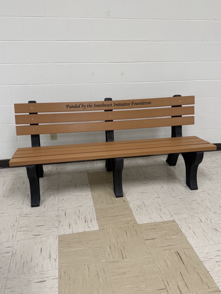 A wooden bench with black legs sits against a white brick wall. The backrest reads, Funded by the Southwest Initiative Foundation. The floor is tiled in beige and gray.