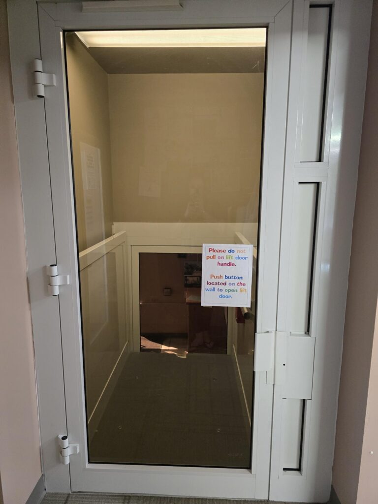 Glass door in a hallway with a sign that says, Please do not pull on side door handle. Push button located on the door. Stairs are visible behind the door.