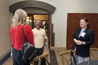 Three people stand and smile while talking in a room with wooden doors. The woman on the left has long blonde hair and a red top, facing two people, one man and one woman, who both have name tags and are standing near a table.