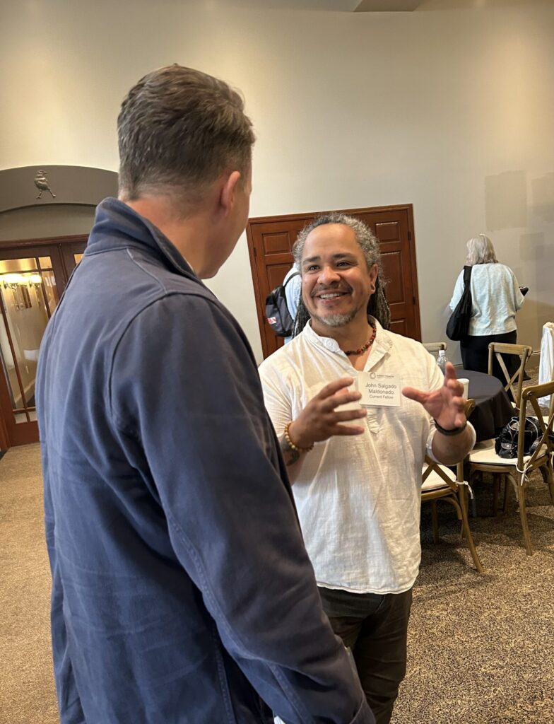 Two men stand and talk at an indoor event. One gestures while speaking and smiles. Behind them, people sit and a TV screen displays a persons name and photo. The setting appears casual and relaxed.