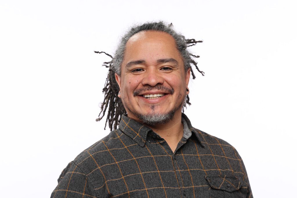 A smiling man with medium-length dreadlocks and a short beard, wearing a plaid shirt, stands against a plain white background.