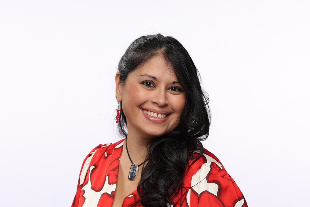 Smiling woman with long dark hair, wearing a red and white patterned top, red earrings, and a black pendant necklace, posed against a plain white background.