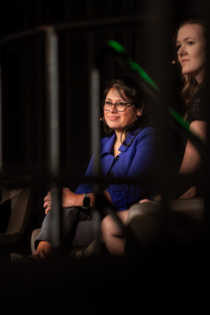 Two women sit side by side on a dimly lit stage, one wearing glasses and a blue blazer, both with microphones. Out-of-focus railings are visible in the foreground.