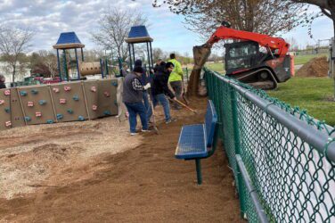 Several people spread dirt along a playground fence with rakes while a small loader vehicle pours more dirt nearby. Playground equipment and benches are visible under a partly cloudy sky.
