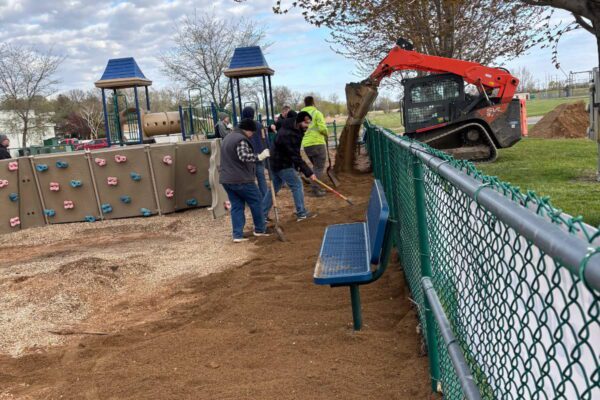 Several people spread dirt along a playground fence with rakes while a small loader vehicle pours more dirt nearby. Playground equipment and benches are visible under a partly cloudy sky.