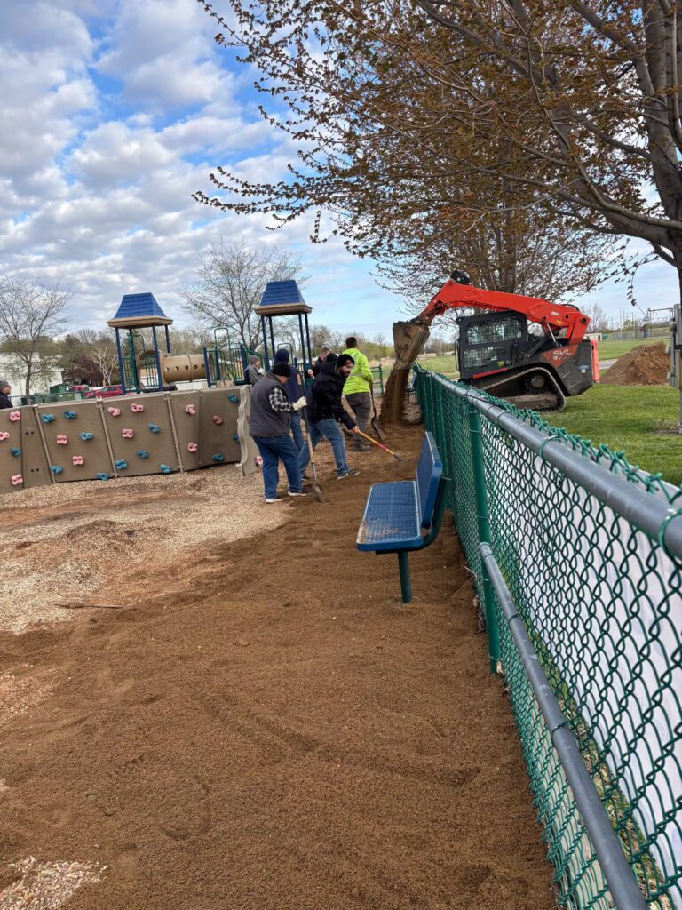 Several people spread dirt along a playground fence with rakes while a small loader vehicle pours more dirt nearby. Playground equipment and benches are visible under a partly cloudy sky.