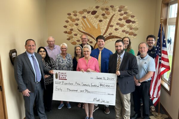 A group of people stand in an office holding a large check for $40,000 made out to the Pipestone Area Community Foundation. An American flag stands in the corner and a tree mural decorates the wall behind them.