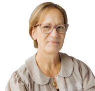 A woman with short, light brown hair and glasses, wearing a beige blouse and a gold necklace, smiles slightly while looking at the camera. The background is plain white.