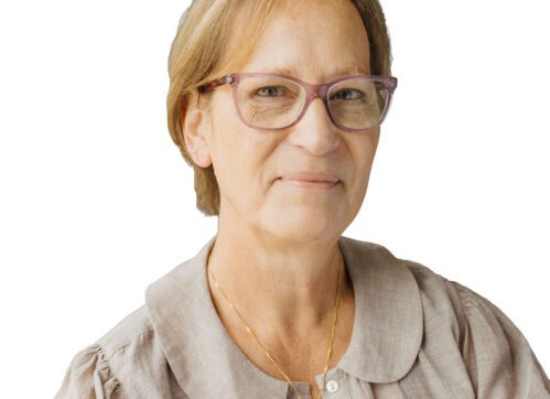 A woman with short, light brown hair and glasses, wearing a beige blouse and a gold necklace, smiles slightly while looking at the camera. The background is plain white.
