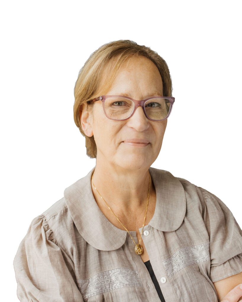 A woman with short, light brown hair and glasses, wearing a beige blouse and a gold necklace, smiles slightly while looking at the camera. The background is plain white.