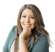 Smiling woman with long, wavy brown hair wearing a green jacket and white top, resting her chin on her hand, posed in front of a white background.
