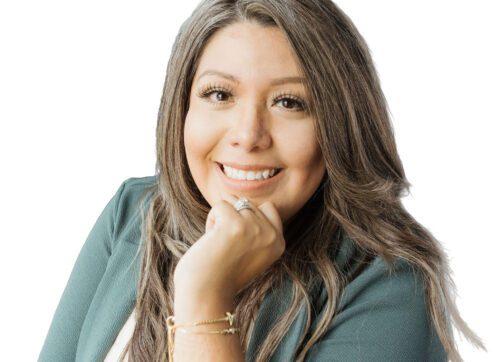 Smiling woman with long, wavy brown hair wearing a green jacket and white top, resting her chin on her hand, posed in front of a white background.