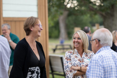 Three adults stand outdoors in conversation, smiling and laughing. One woman wears glasses and a black dress, another wears a floral blouse, and a man with white hair wears glasses and a blue-striped shirt. Trees are visible in the background.